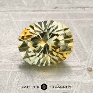 A 2.15-carat mint green-yellow particolored Montana sapphire (heated) is displayed on a textured silver surface with the text “EARTH’S TREASURY” below.