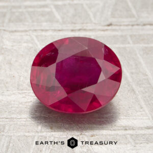 A 2.07-carat Mozambique ruby (heated), cut in an oval shape, is displayed on a light gray textured surface with “Earth’s Treasury” text below.