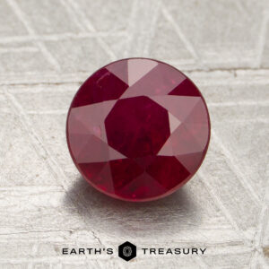 A 1.86-carat Mozambique ruby (heated), round cut, is displayed on a textured surface with the text “EARTH’S TREASURY” and a hexagonal logo visible at the bottom of the image.