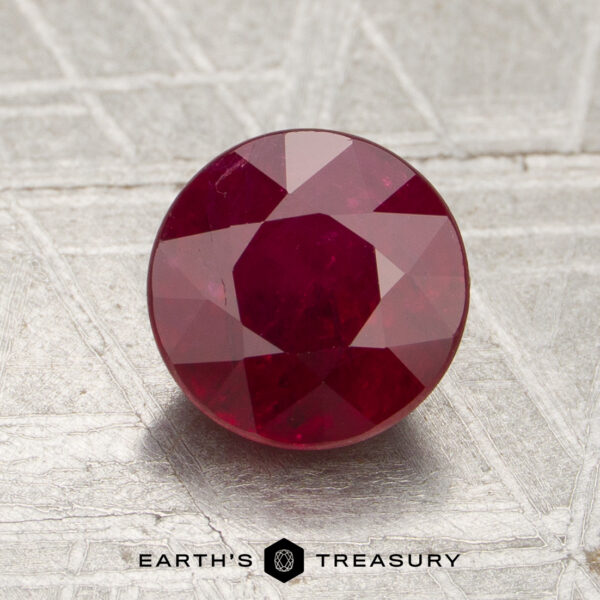 1.86-Carat Mozambique Ruby (Heated) A 1.86-carat Mozambique ruby (heated), round cut, is displayed on a textured surface with the text “EARTH’S TREASURY” and a hexagonal logo visible at the bottom of the image.