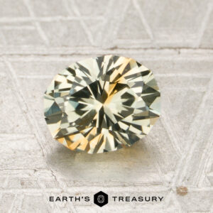 A 1.80-carat pale green-orange particolored Montana sapphire (heated) with an oval faceted cut is showcased on a textured gray surface, accompanied by the “EARTH’S TREASURY” text and a hexagon logo at the bottom.