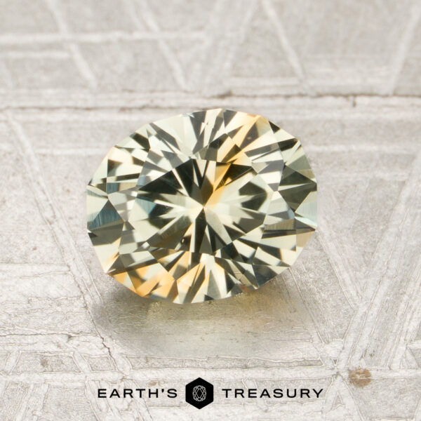 1.80-Carat Pale Green-Orange Particolored Montana Sapphire (Heated) A 1.80-carat pale green-orange particolored Montana sapphire (heated) with an oval faceted cut is showcased on a textured gray surface, accompanied by the “EARTH’S TREASURY” text and a hexagon logo at the bottom.