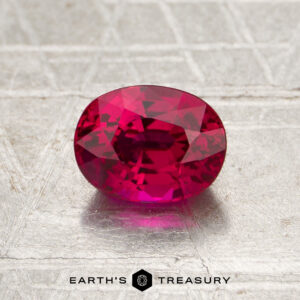 A 1.20-carat Mozambique ruby (heated) in an oval cut is showcased on a textured, metallic surface with the text “EARTH’S TREASURY” at the bottom.