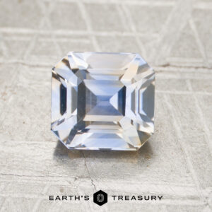 A rectangular, emerald-cut 2.05-carat yellow-sky blue particolored Ceylon sapphire (heated) rests on a textured surface, with the Earth’s Treasury logo and name visible at the bottom of the image.