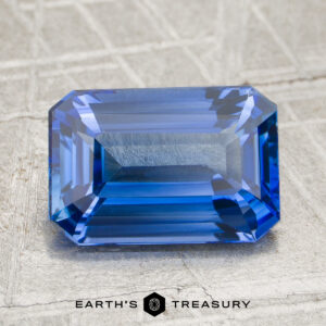 A 3.00-carat rich cornflower blue Ceylon sapphire (heated) with an emerald cut lies on a textured metallic surface, with “EARTH’S TREASURY” text and logo beneath it.