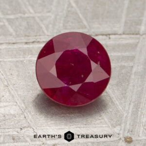 A round, faceted 1.55-carat Mozambique Ruby (Heated) displayed on a textured gray surface with the text “EARTH’S TREASURY” below the stone.