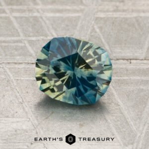 A 1.37-carat blue-yellow particolored Montana sapphire, heated and brilliantly cut in an oval shape, rests on a textured surface. The text “EARTH’S TREASURY” appears at the bottom of the image.