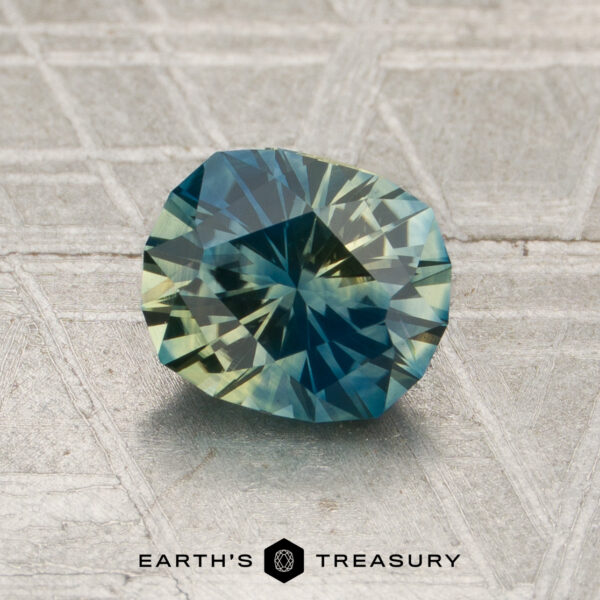 1.37-Carat Blue-Yellow Particolored Montana Sapphire (Heated) A 1.37-carat blue-yellow particolored Montana sapphire, heated and brilliantly cut in an oval shape, rests on a textured surface. The text “EARTH’S TREASURY” appears at the bottom of the image.