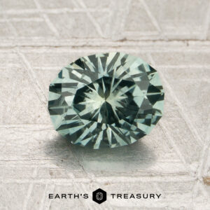 A round, faceted 1.70-carat mint green Montana sapphire (heated) rests on a textured metallic surface. The logo and text “EARTH’S TREASURY” are at the bottom of the image.