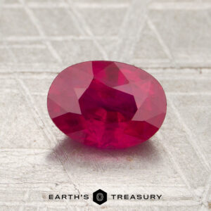 A 1.28-carat Burmese ruby (heated), oval in shape and vivid red, is showcased on a textured surface with the “Earth’s Treasury” logo and text visible at the bottom of the image.