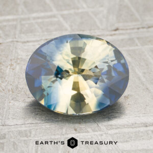 A 2.56-carat yellow-blue particolored sapphire (heated) with a round faceted cut rests on a textured surface, while the logo and text “EARTH’S TREASURY” appear at the bottom of the image.