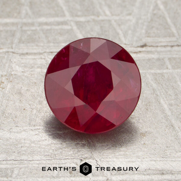 1.51-Carat Mozambique Ruby (Heated) A 1.51-carat round-cut Mozambique ruby (heated) displayed on a textured surface, with “Earth’s Treasury” and a hexagonal logo beneath the gemstone.