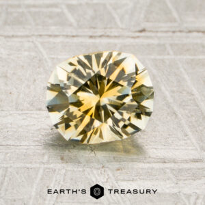 A 1.34-carat yellow-colorless particolored Montana sapphire (heated) is displayed on a textured surface, with the text “Earth’s Treasury” and a hexagon logo beneath it.