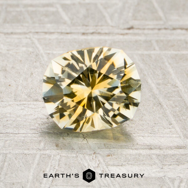 A 1.34-carat yellow-colorless particolored Montana sapphire (heated) is displayed on a textured surface, with the text “Earth’s Treasury” and a hexagon logo beneath it.