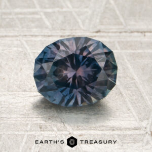 A 1.69-carat steel blue-purple particolored Montana sapphire (heated), faceted in an oval shape, rests on a textured metallic surface with the Earth’s Treasury logo below.