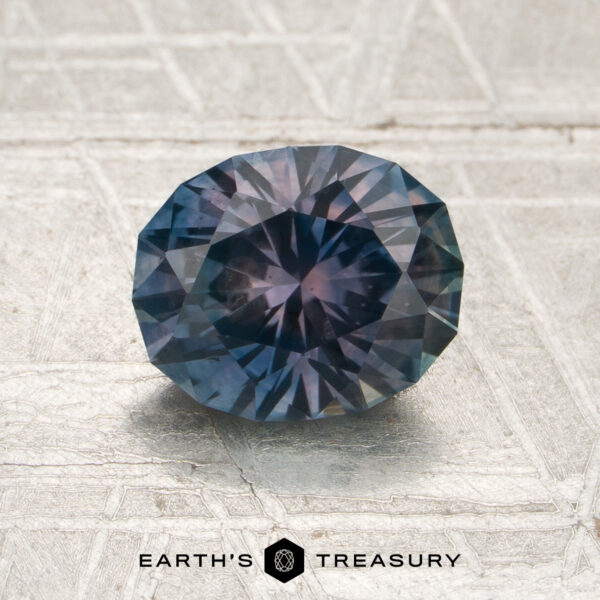 1.69-Carat Steel Blue-Purple Particolored Montana Sapphire (Heated) A 1.69-carat steel blue-purple particolored Montana sapphire (heated), faceted in an oval shape, rests on a textured metallic surface with the Earth’s Treasury logo below.