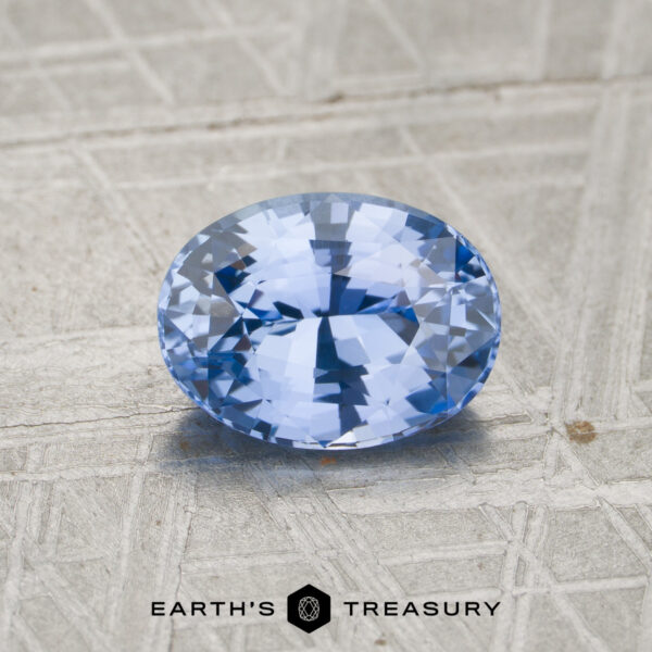 2.21-Carat Sky Blue Ceylon Sapphire (Heated) A 2.21-carat sky blue to violet color-change Ceylon sapphire (heat-treated), cut in an oval shape, is showcased on a textured, light-colored surface with the “Earth’s Treasury” logo and text at the bottom.