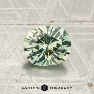 A 1.27-carat mint green Montana sapphire (heated) rests on a textured surface, with the words “EARTH’S TREASURY” and a hexagon logo displayed below it.