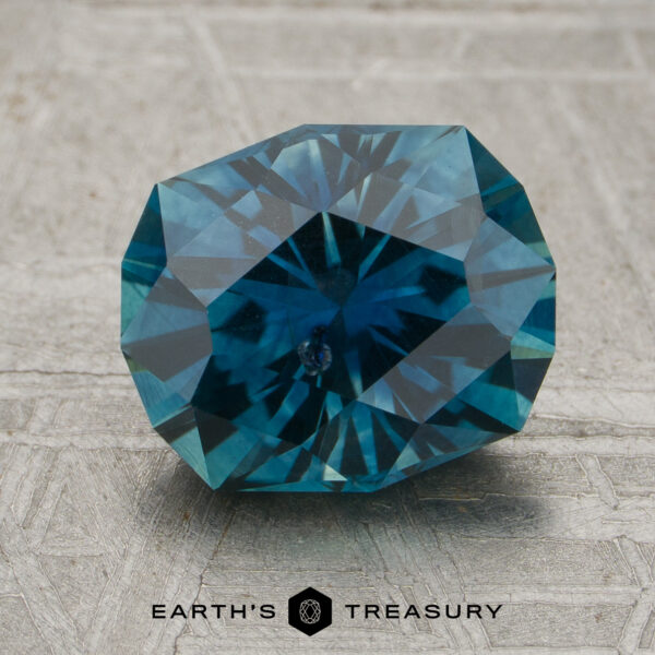 2.46-Carat Peacock Blue Montana Sapphire (Heated) A 2.46-carat Peacock Blue Montana Sapphire (heated) with a geometric cut is displayed on a textured gray surface, with the “Earth’s Treasury” name and hexagon logo shown at the bottom of the image.