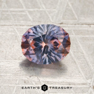A 1.61-carat purple-orange particolored Montana sapphire (heated) with striking blue and orange hues rests on a textured metallic surface, while the “EARTH’S TREASURY” logo and text appear at the bottom.