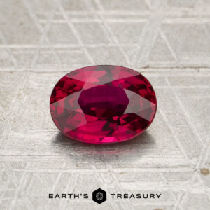 A 2.12-carat heated Madagascar ruby, cut in a faceted oval shape, rests on a textured metallic surface above the “EARTH’S TREASURY” name and geometric logo.