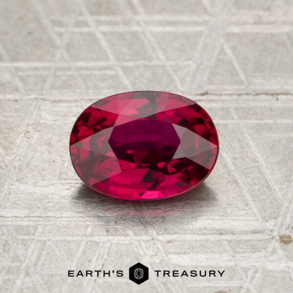 A 2.12-carat heated Madagascar ruby, cut in a faceted oval shape, rests on a textured metallic surface above the “EARTH’S TREASURY” name and geometric logo.