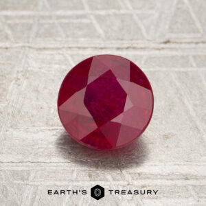 A round, faceted 1.42-carat Mozambique ruby (heated) sits on a textured surface, with the text “Earth’s Treasury” and a logo below.