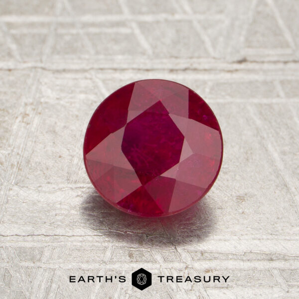 A round, faceted 1.42-carat Mozambique ruby (heated) sits on a textured surface, with the text “Earth’s Treasury” and a logo below.