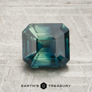 A 2.04-carat green-teal particolored Madagascar sapphire (heated), cut in a rectangular emerald shape, is displayed on a textured light surface with the Earth’s Treasury logo beneath it.