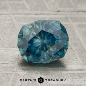 A 1.74-carat teal Montana sapphire (heated) rests on a textured gray surface with the “Earth’s Treasury” logo and text below it.