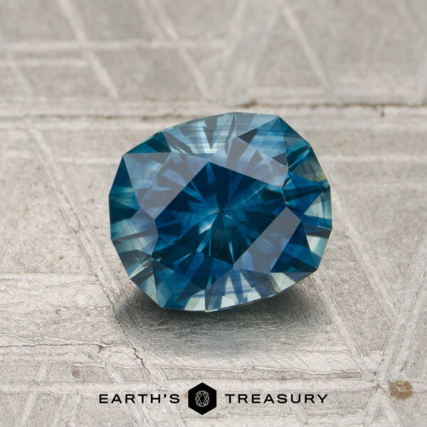 1.46-Carat Teal Montana Sapphire (Heated) A 1.46-carat heated Montana sapphire is displayed on a textured, silvery surface with the words “Earth’s Treasury” printed below.