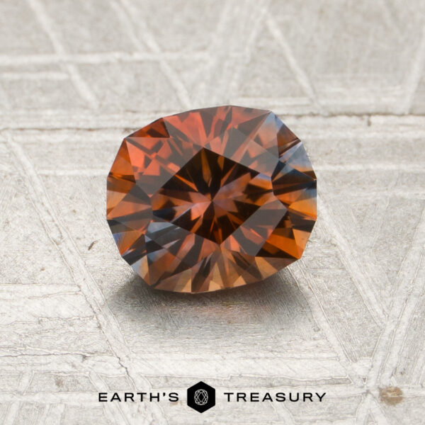A 1.24-carat burnt orange-gray particolored Montana sapphire (heated) with an oval cut and multiple facets is displayed on a textured gray surface, with “EARTH’S TREASURY” visible at the bottom.
