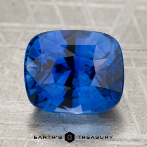 A 4.39-carat Royal Blue Ceylon sapphire (heated) with a cushion cut is displayed on a textured gray surface, with the words “Earth’s Treasury” at the bottom.