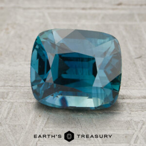 A rectangular cushion-cut 3.21-carat rich teal Madagascar sapphire (heated) rests on a textured gray surface with the words “Earth’s Treasury” at the bottom.