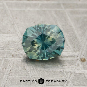 A 0.98-carat teal-yellow particolored Montana sapphire (heated) with a faceted oval shape rests on a textured gray surface, with “Earth’s Treasury” displayed beneath it.