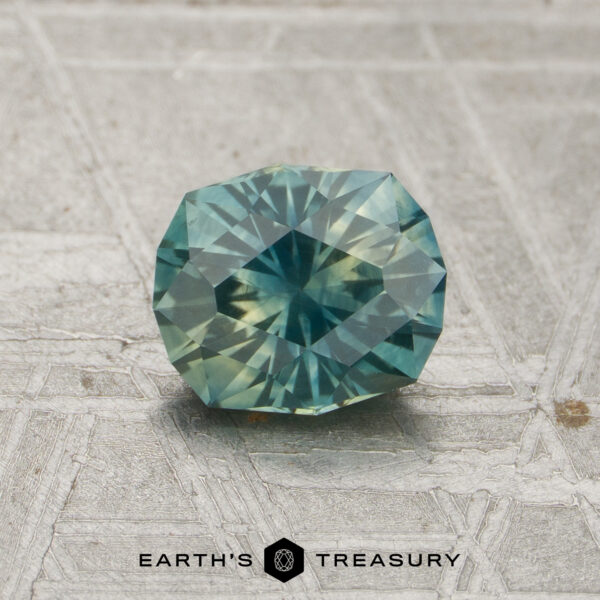 A 0.98-carat teal-yellow particolored Montana sapphire (heated) with a faceted oval shape rests on a textured gray surface, with “Earth’s Treasury” displayed beneath it.