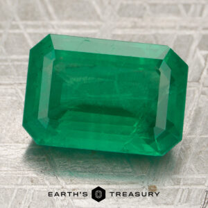 A 6.52-carat Zambian emerald, cut in a rectangular shape, rests on a textured metallic surface with the text “Earth’s Treasury” at the bottom.