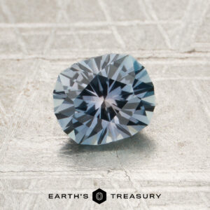 A round, faceted 1.18-carat steely light blue Montana sapphire (heated) rests on a textured surface. The logo and text “EARTH’S TREASURY” appear at the bottom of the image.