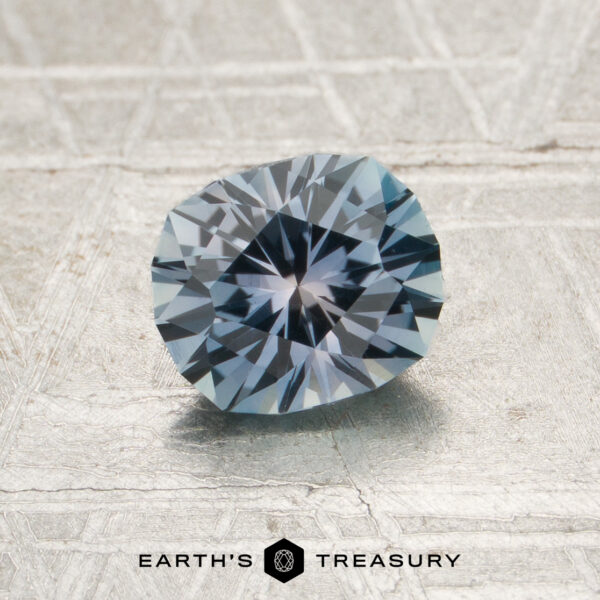 1.18-Carat Steely Light Blue Montana Sapphire (Heated) A round, faceted 1.18-carat steely light blue Montana sapphire (heated) rests on a textured surface. The logo and text “EARTH’S TREASURY” appear at the bottom of the image.