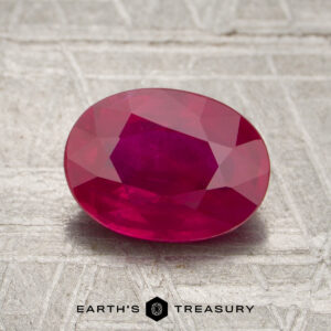 A 1.97-carat Burmese ruby (heated), cut in an oval faceted shape, is displayed on a textured gray surface. The text “EARTH’S TREASURY” appears at the bottom of the image.