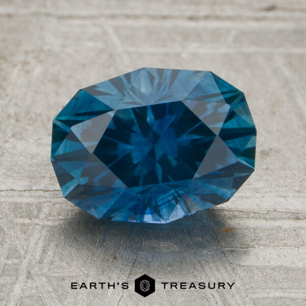 2.34-Carat Rich Teal Blue Montana Sapphire (Heated) A 2.34-carat rich teal blue Montana sapphire (heated) is displayed on a textured surface, with the words “Earth’s Treasury” and a hexagonal logo below it.