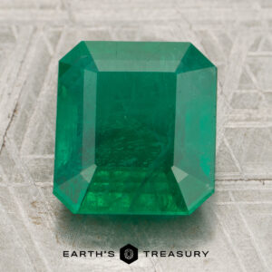 A 10.11-carat Zambian emerald, cut in a rectangular shape, displayed on a textured gray surface with the words “Earth’s Treasury” below the gemstone.
