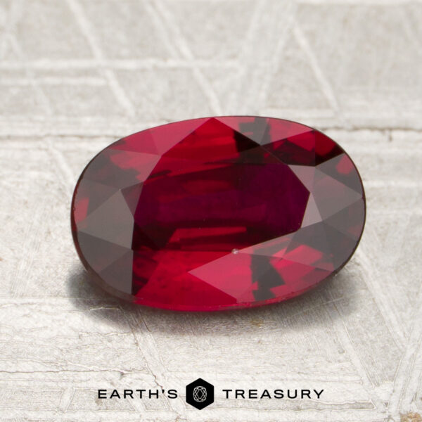 2.01-Carat Mozambique Ruby (Heated) A 2.01-carat heated Mozambique ruby in an oval cut is displayed on a textured surface, with “Earth’s Treasury” appearing at the bottom of the image.