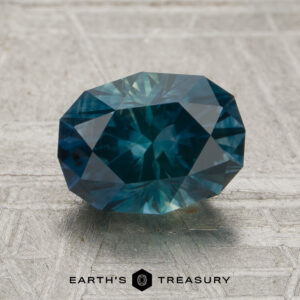 A 1.98-carat deep blue-green Montana sapphire (heated) in a faceted oval cut is showcased on a textured gray surface featuring the “Earth’s Treasury” logo at the bottom.