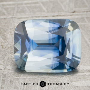 A 3.55-carat colorless-blue particolored Ceylon sapphire with facets rests on a textured gray surface. The logo and text “Earth’s Treasury” appear at the bottom of the image.