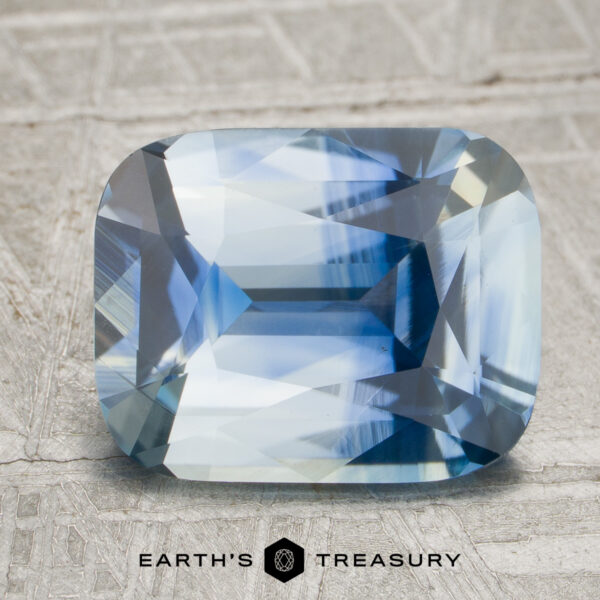 3.55-Carat Colorless-Blue Particolored Ceylon Sapphire (Unheated) A 3.55-carat colorless-blue particolored Ceylon sapphire with facets rests on a textured gray surface. The logo and text “Earth’s Treasury” appear at the bottom of the image.