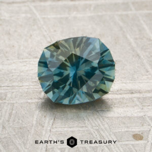 A faceted oval 1.17-carat blue-yellow particolored Montana sapphire (heated) is displayed on a textured surface. The logo and text “EARTH’S TREASURY” appear at the bottom of the image.