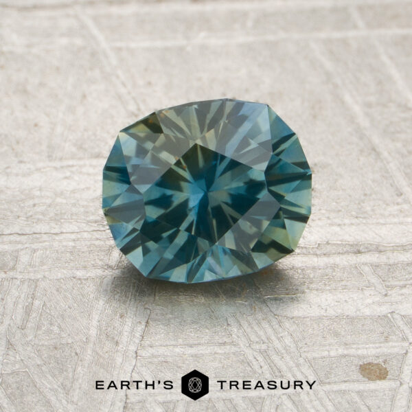1.17-Carat Blue-Yellow Particolored Montana Sapphire (Heated) A faceted oval 1.17-carat blue-yellow particolored Montana sapphire (heated) is displayed on a textured surface. The logo and text “EARTH’S TREASURY” appear at the bottom of the image.