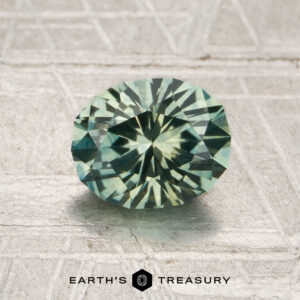 A 1.52-carat rich mint green Montana sapphire (heated) rests on a textured metallic surface, with the text “Earth’s Treasury” below the stone.