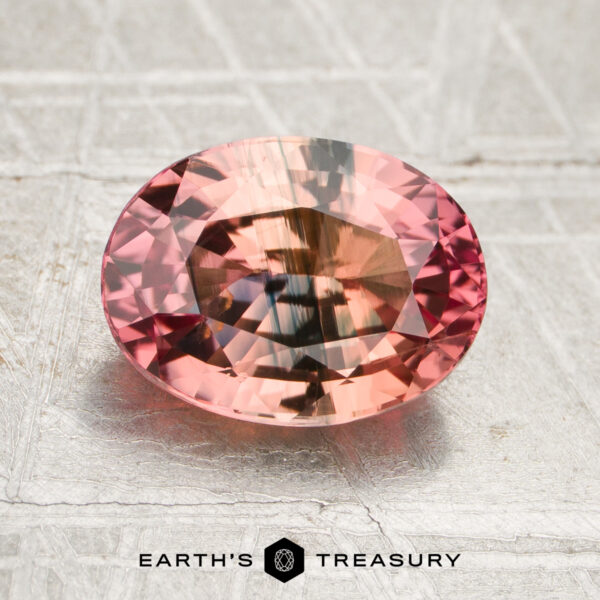 2.55-Carat Peach Pink-Teal Particolored Ceylon Sapphire (Heated) A 2.55-carat peach pink-teal particolored sapphire (heated), oval-cut with reflective facets, rests on a textured metallic surface; the “Earth’s Treasury” logo and text are visible at the bottom.